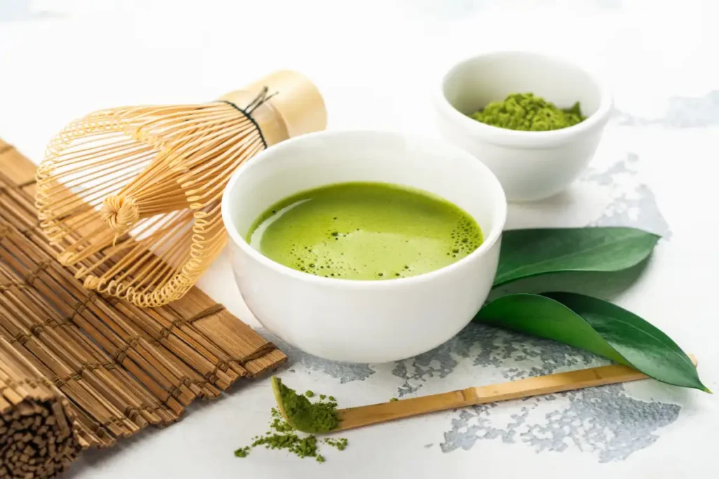 A glass cup of green matcha tea next to a separate bowl of matcha powder, both showcasing the vibrant green color against a light background.