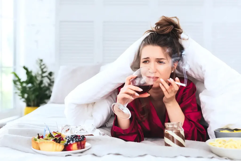 Heartbroken woman lying in bed, smoking a cigarette and drinking wine, lost in deep thoughts.