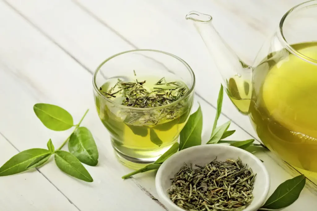 A glass of herbal tea next to a glass teapot filled with green tea, with fresh green tea leaves in a cup ready to be brewed.