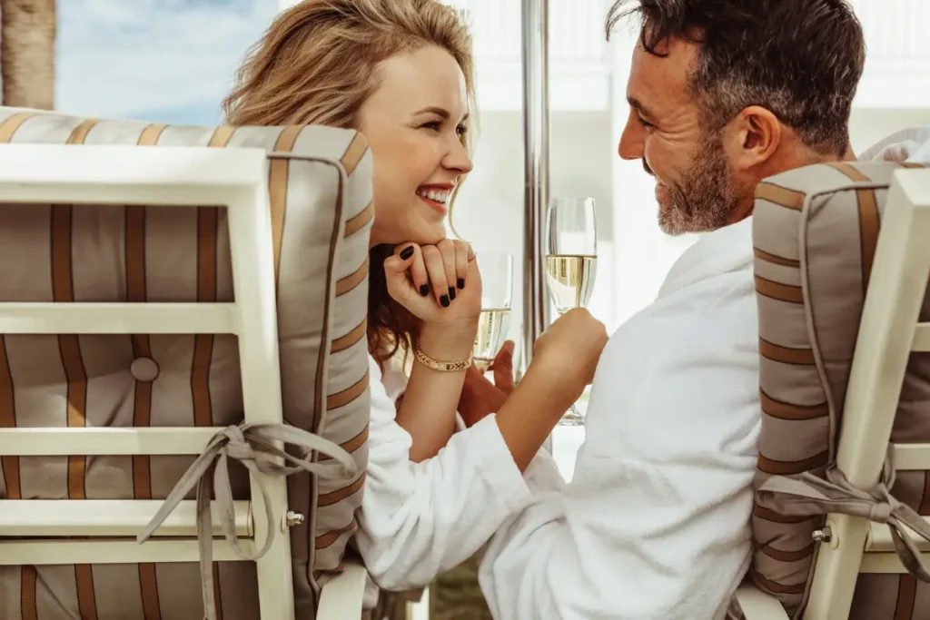 Loving couple sitting in chairs, smiling at each other while drinking glasses of white wine, enjoying a cozy moment together.