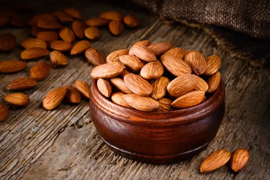 Raw almonds scattered on a wooden table, with some placed in a rustic wooden bowl, adding to the natural feel.
