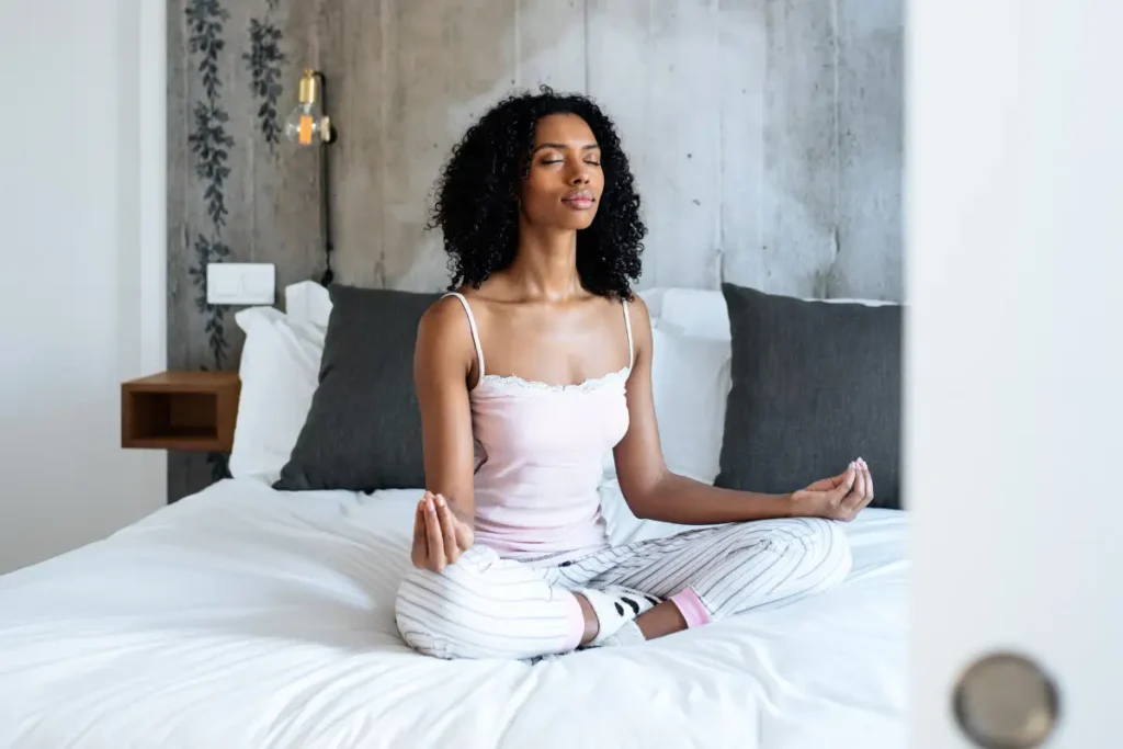 A peaceful black woman meditating in Sukhasana (Easy Pose) on her bed, practicing mindfulness and relaxation.