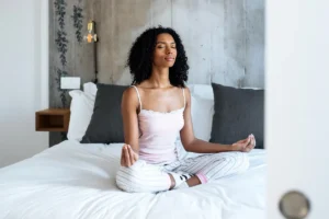 A peaceful black woman meditating in Sukhasana (Easy Pose) on her bed, practicing mindfulness and relaxation.