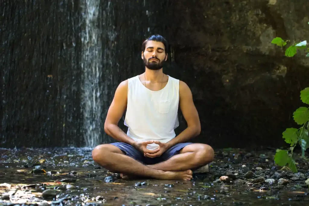 A man meditating peacefully in nature, with a small waterfall flowing nearby, surrounded by greenery and tranquil sounds.