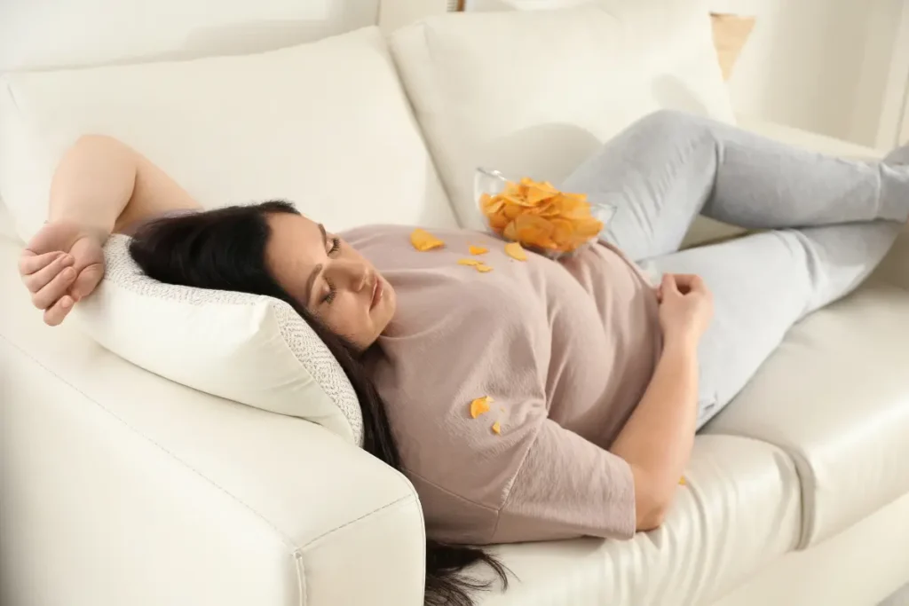 Plus-size woman falling asleep while eating chips, representing eating habits.
