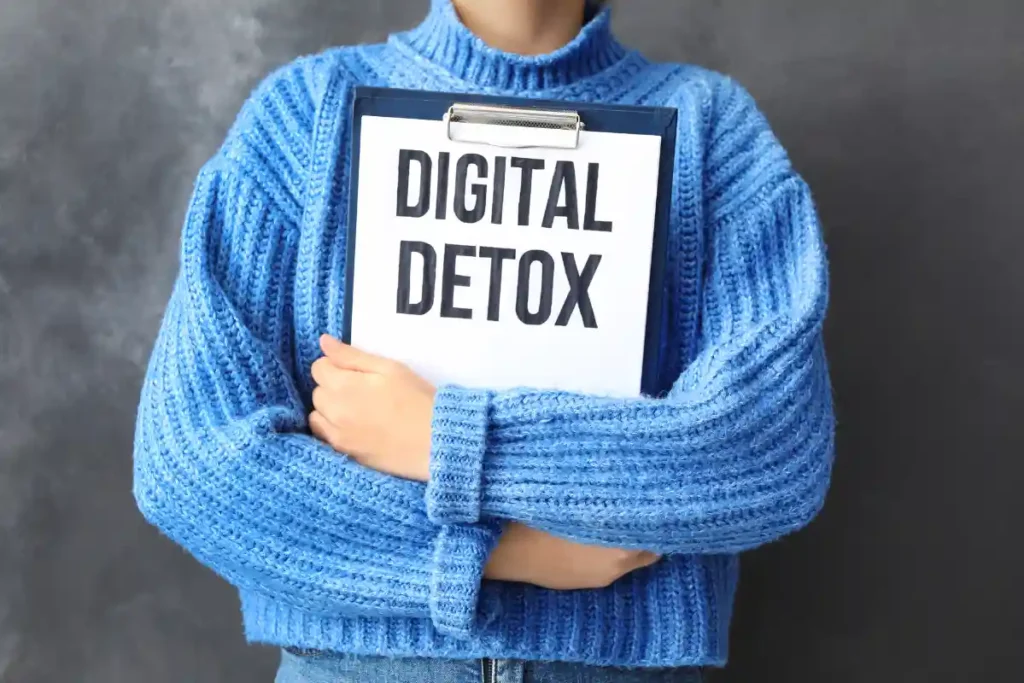 "Person leaning against a wall, holding an open office folder against their chest with the message 'Digital Detox' written on a sheet.