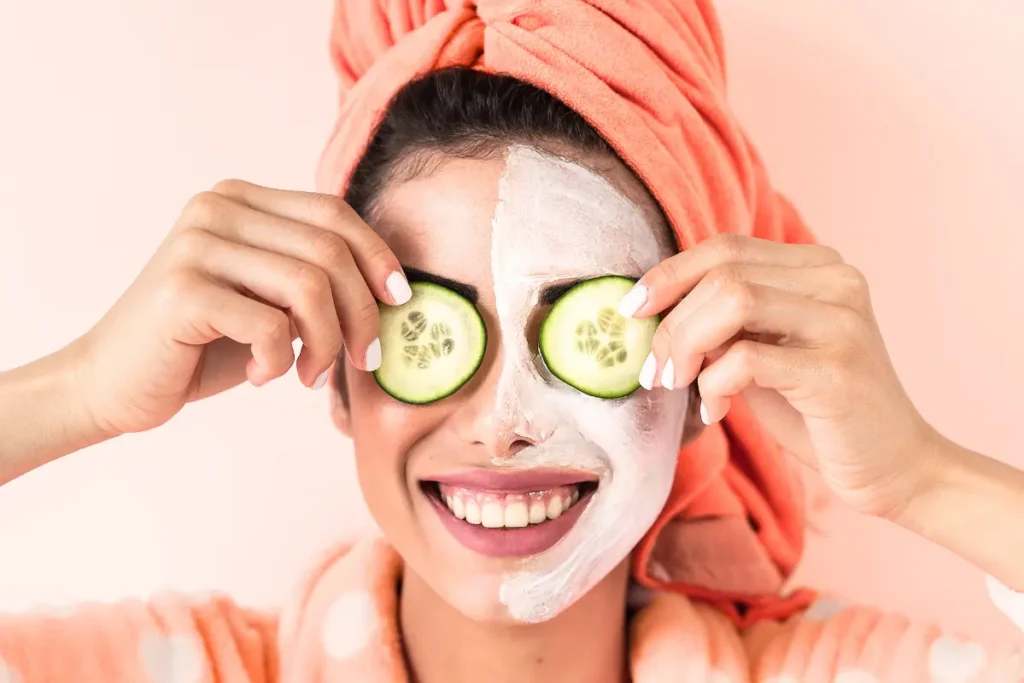 Woman at home following a skincare routine, with one half of her face covered in a mask and the other bare, while cucumber slices rest on each of her eyes.