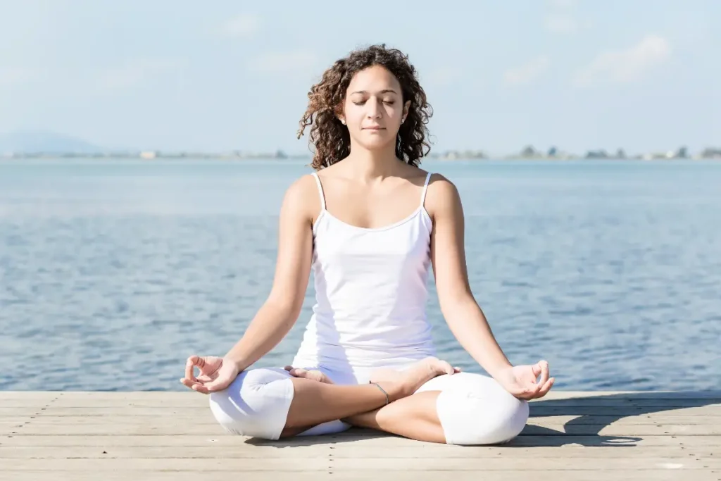 A woman practicing Padmasana (Lotus Pose) on a sidewalk with the sea in the background, in a peaceful and meditative posture, with a calm outdoor setting.