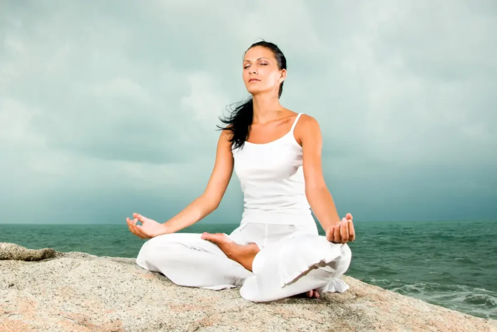 A woman practicing meditation in a peaceful pose on a rocky beach, surrounded by the ocean and natural scenery.