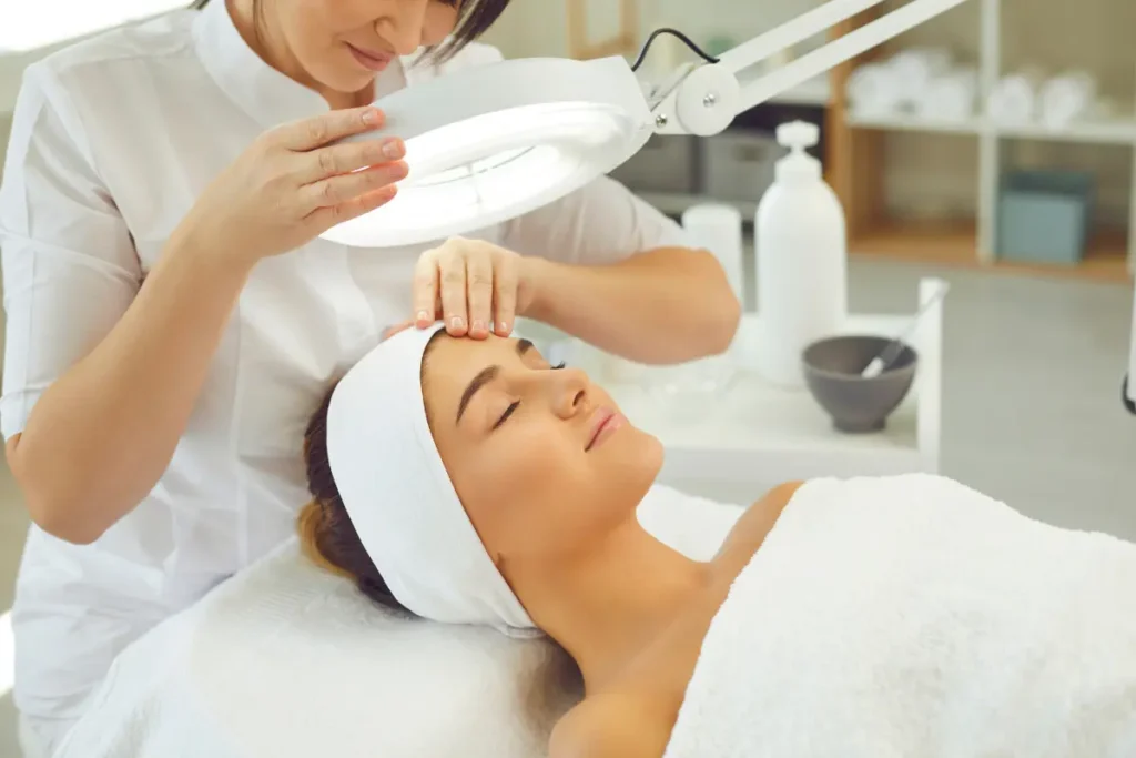 Woman lying on a treatment bed in a dermatologist's office, while the dermatologist uses a magnifying lamp to examine her skin for wrinkles and other concerns.