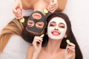 Two women at home enjoying a skincare routine, wearing face masks and holding cucumber slices in each hand.
