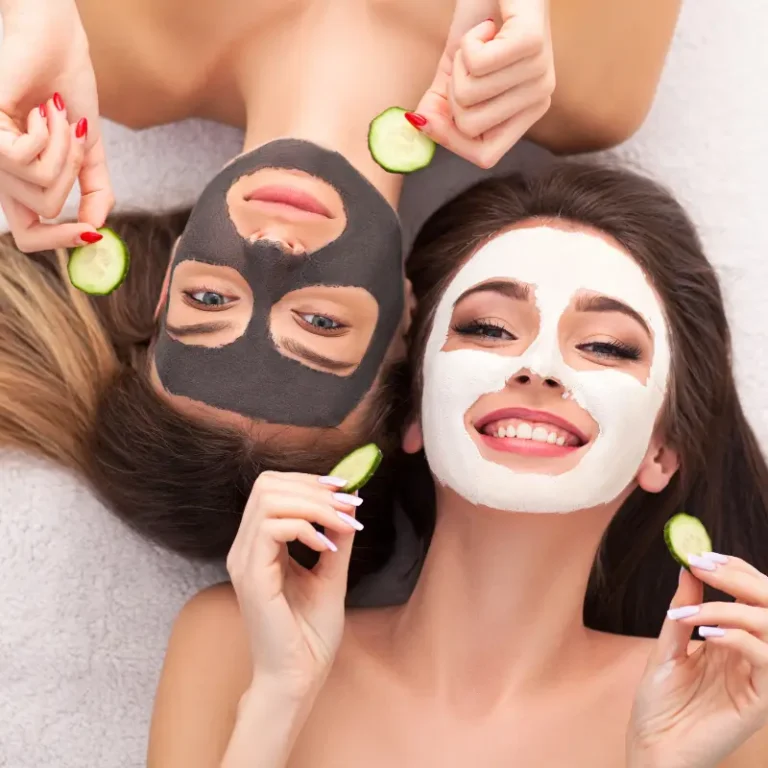 Two women at home enjoying a skincare routine, wearing face masks and holding cucumber slices in each hand.
